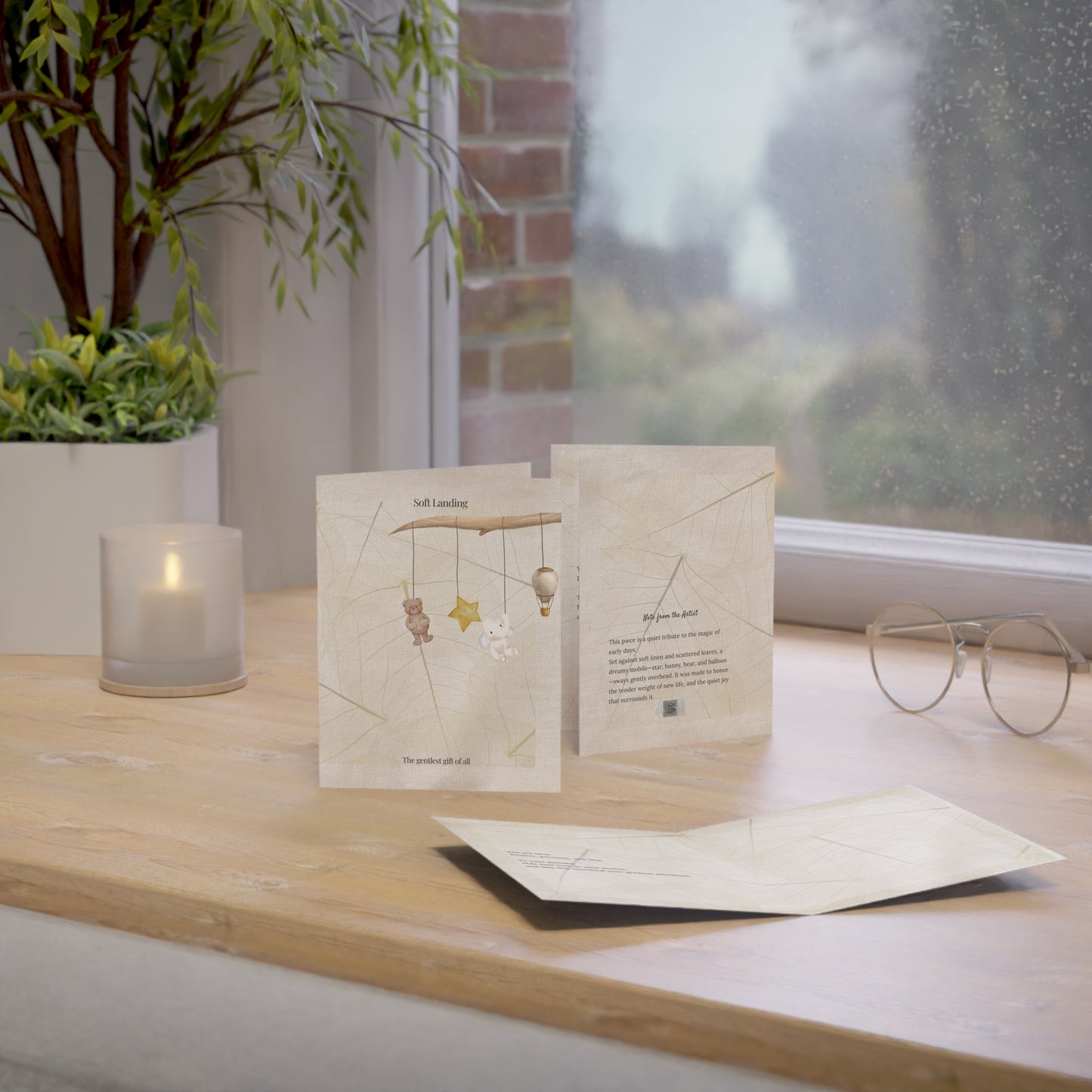 Soft Landing baby card displayed in three views—front cover, back with artist note, and matching envelope—styled by a window with soft daylight, neutral tones, and gentle textures. Perfect for baby arrival or new parent gifts.