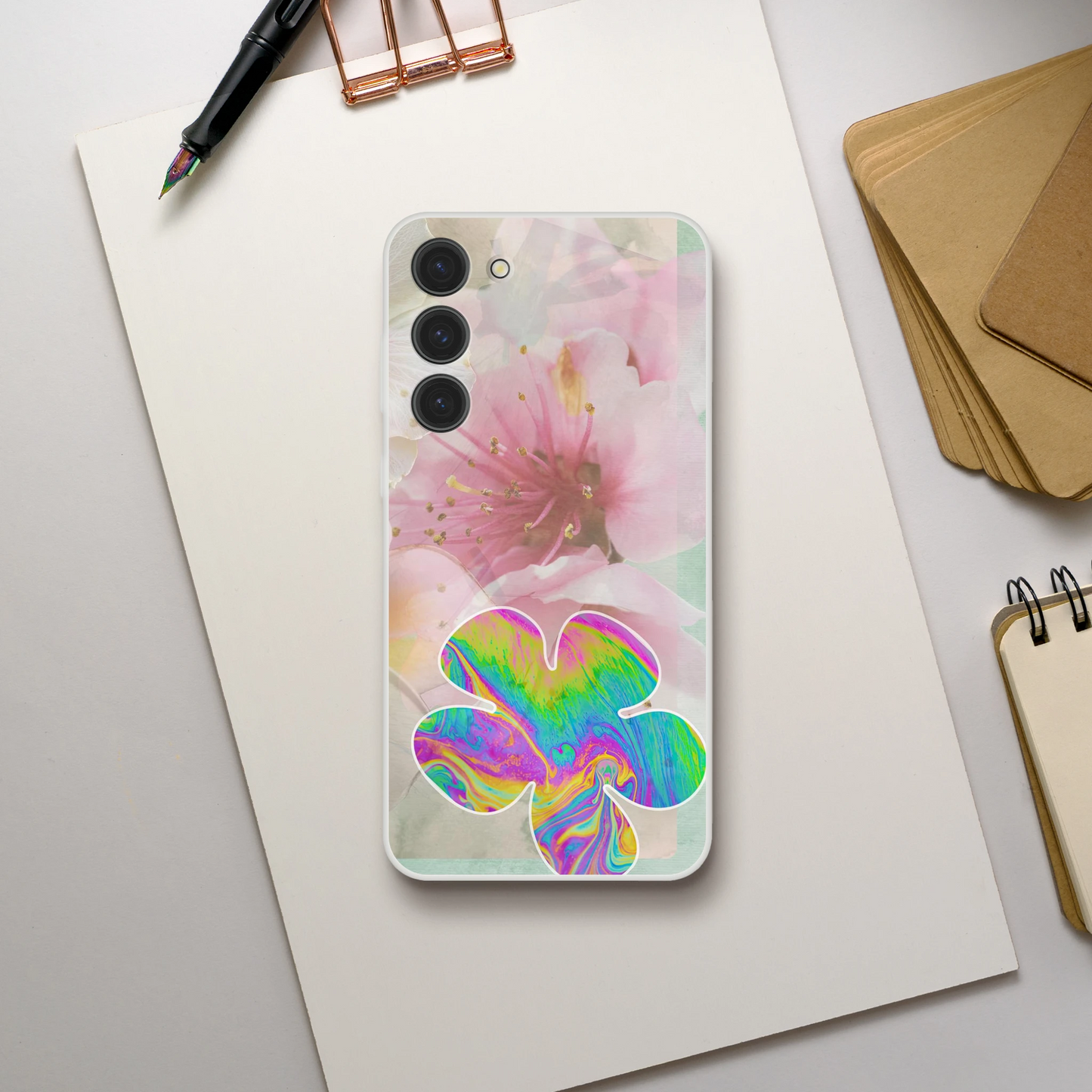 Samsung Galaxy S23 Plus case on a minimalist desk setup with stationery and kraft paper, featuring a transparent background with soft pink cherry blossoms and a bold psychedelic flower for spring style.
