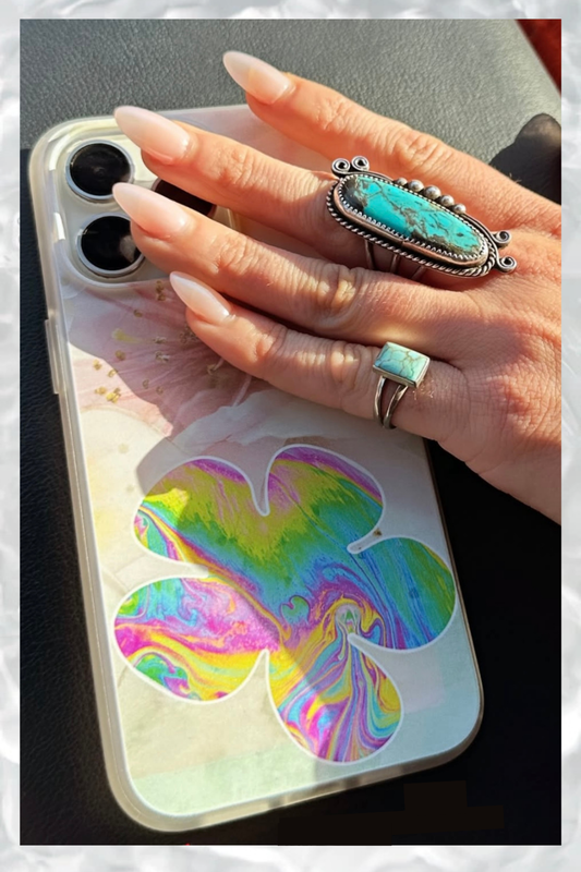 Hand with turquoise statement rings holding a phone with a holographic flower case, glowing in warm sunset light on a textured surface.
