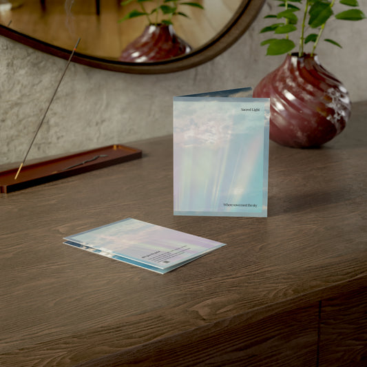 a dreamy wedding card titled "sacred light" displayed upright and flat on a wooden desk besides a red glass vase and incense. The card design features a soft sky with pastel beams of light. 