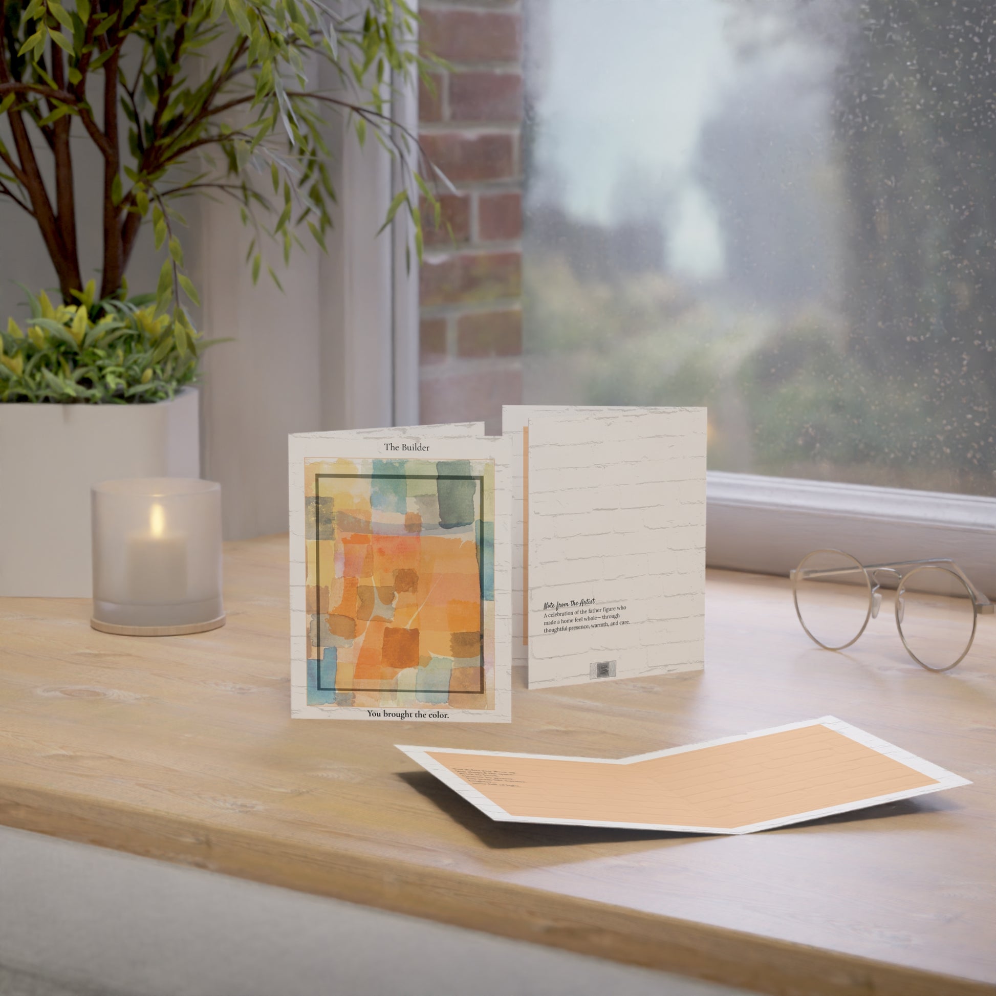 A minimalist art greeting card titled “The Builder,” resting on a wooden desk in natural light. The card features an abstract watercolor design in orange and golden tones, set against a white brick background. An original Father’s Day card for dads, stepdads, or chosen family who bring warmth into everyday life.
