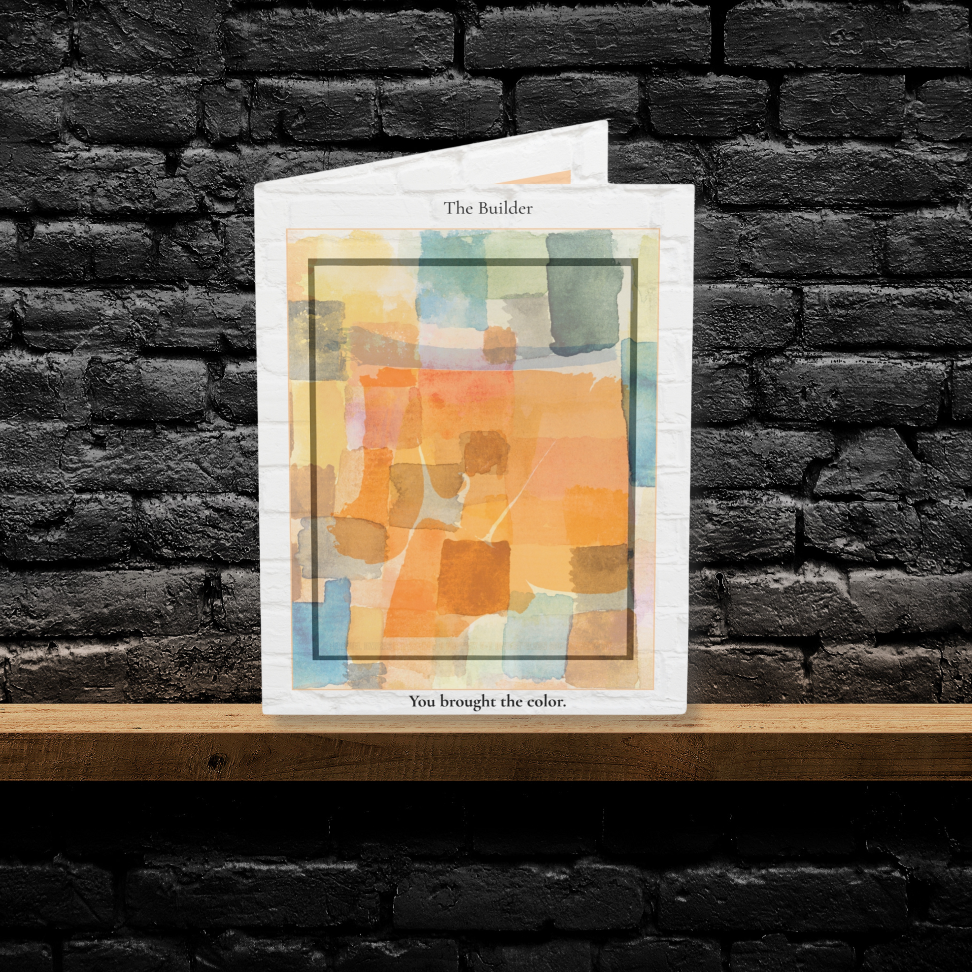 A folded A6 art greeting card titled “The Builder,” featuring an abstract watercolor design in warm orange, yellow, and soft blue tones. Displayed upright on a wooden shelf against a black brick wall. A minimalist greeting card perfect for Father’s Day, appreciation, or gifting to a stepdad or mentor.
